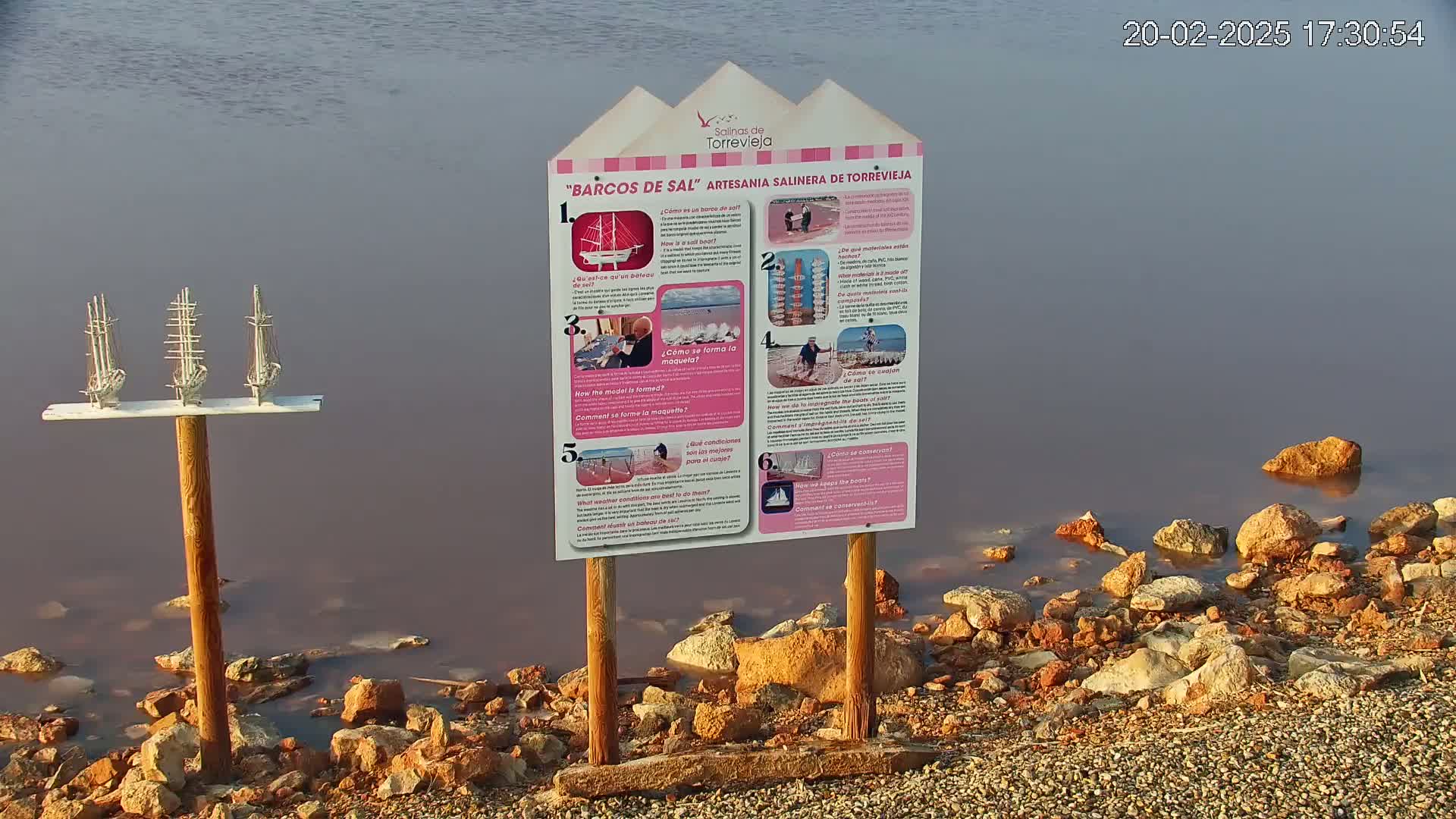 Three small model sailboats stand on a post by a calm body of pinkish water, next to a sign with illustrations and text.