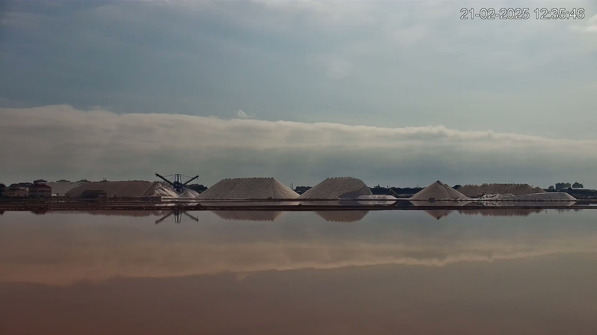 A calm body of water reflects a landscape of several large piles of salt and industrial machinery under a mostly cloudy sky.