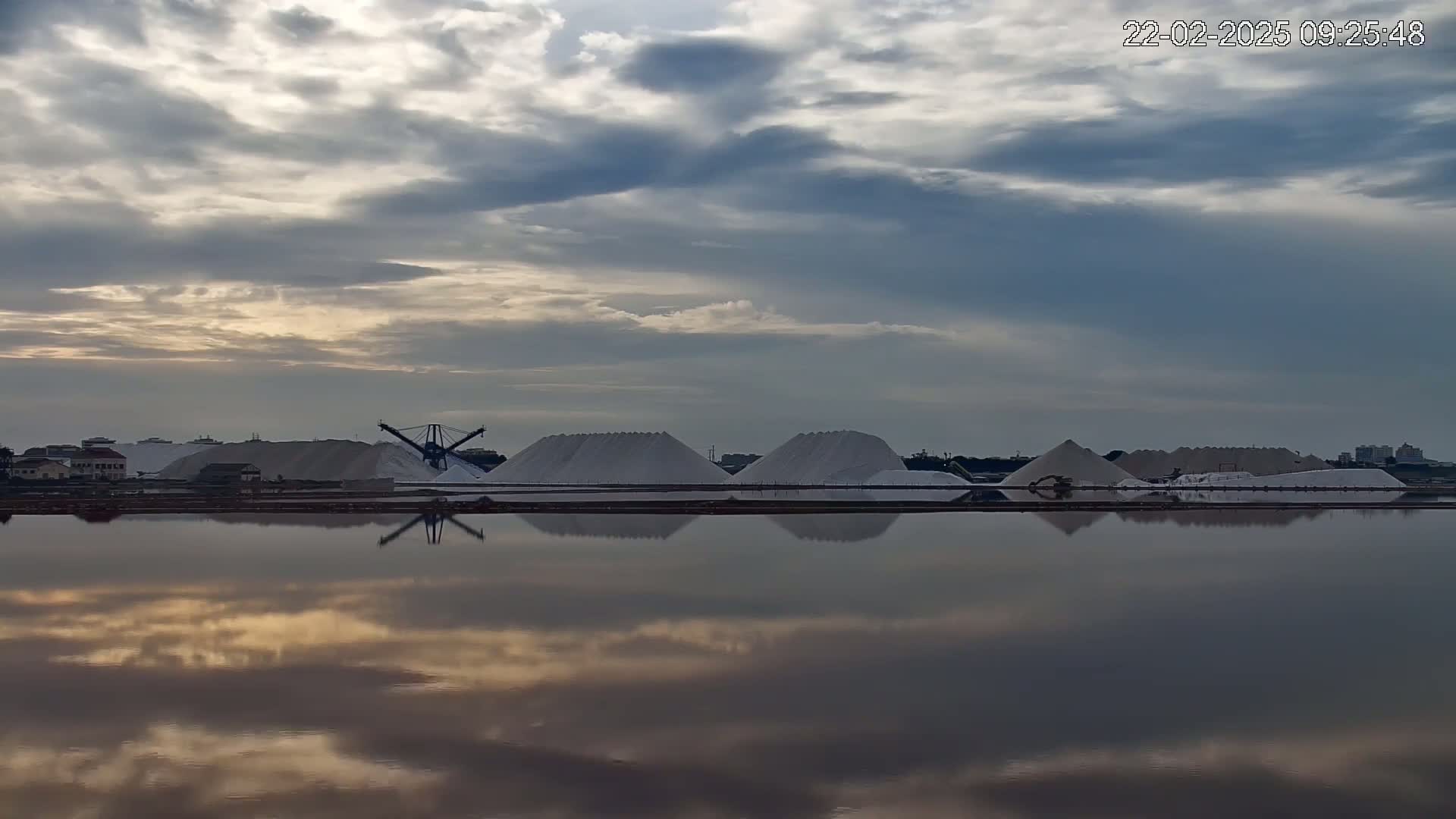Several large piles of white salt are situated beside a calm body of water that reflects the cloudy sky.