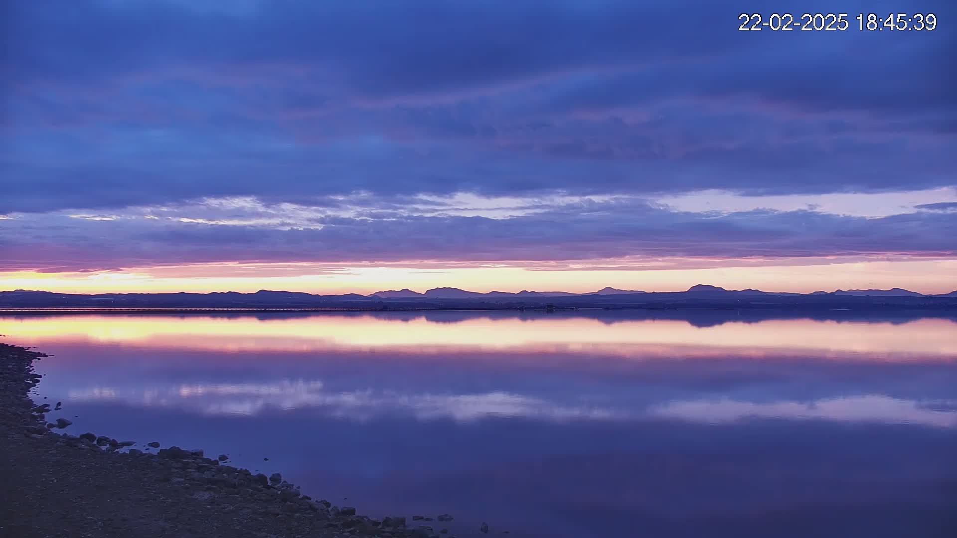 A calm body of water reflects a sunset sky and distant mountains under a partly cloudy sky.