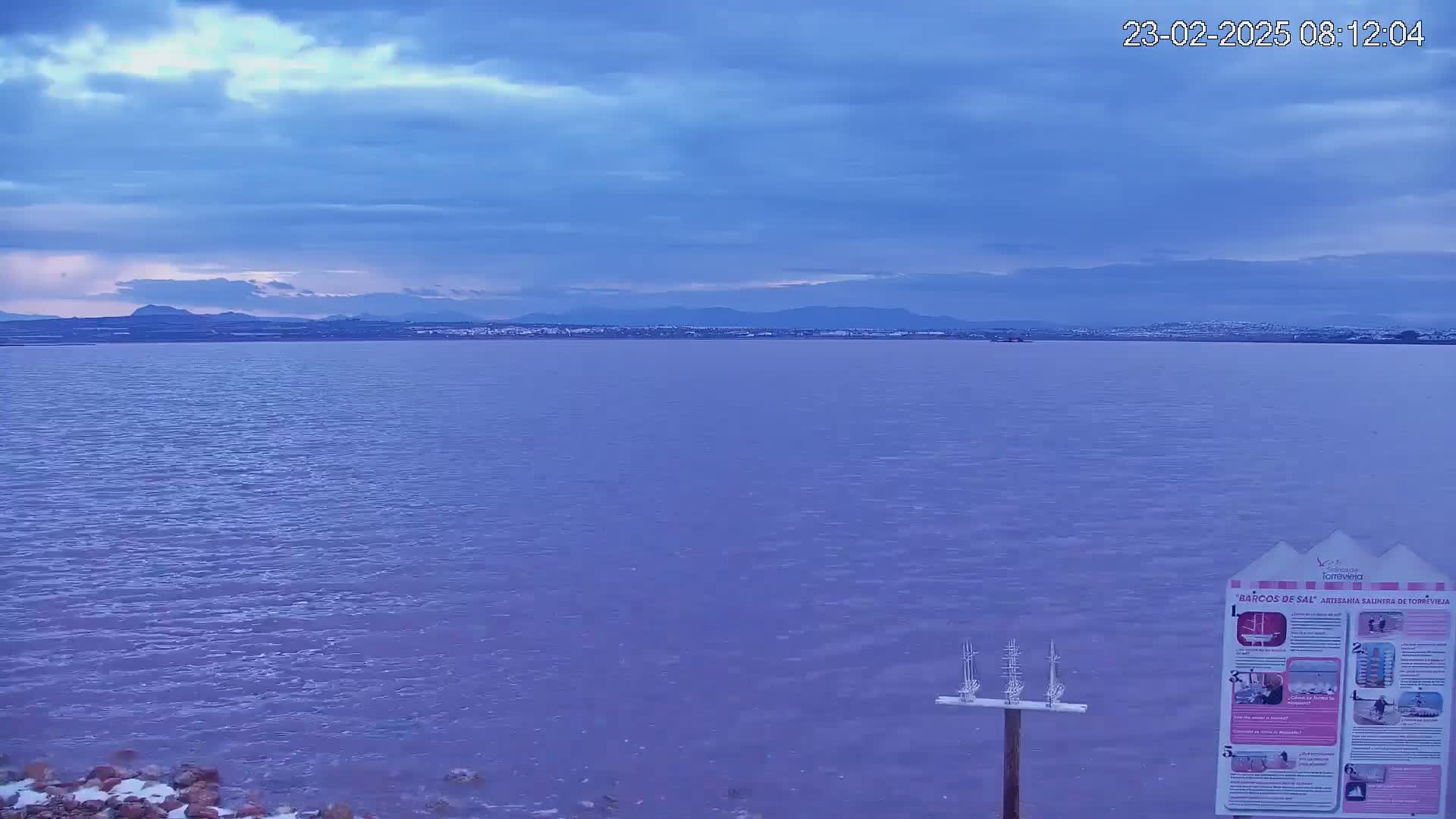 A calm body of water stretches towards a distant mountain range under a cloudy, dusky blue sky.