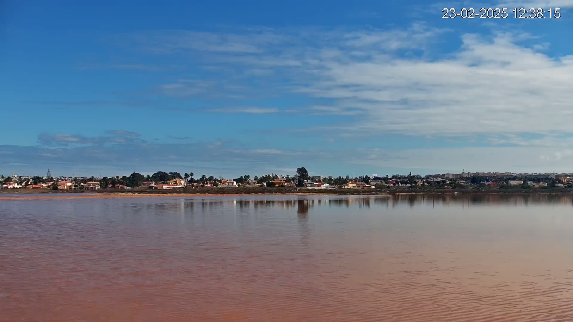 A calm, pinkish-red lake reflects a distant line of houses and buildings under a mostly clear, blue sky with scattered clouds.