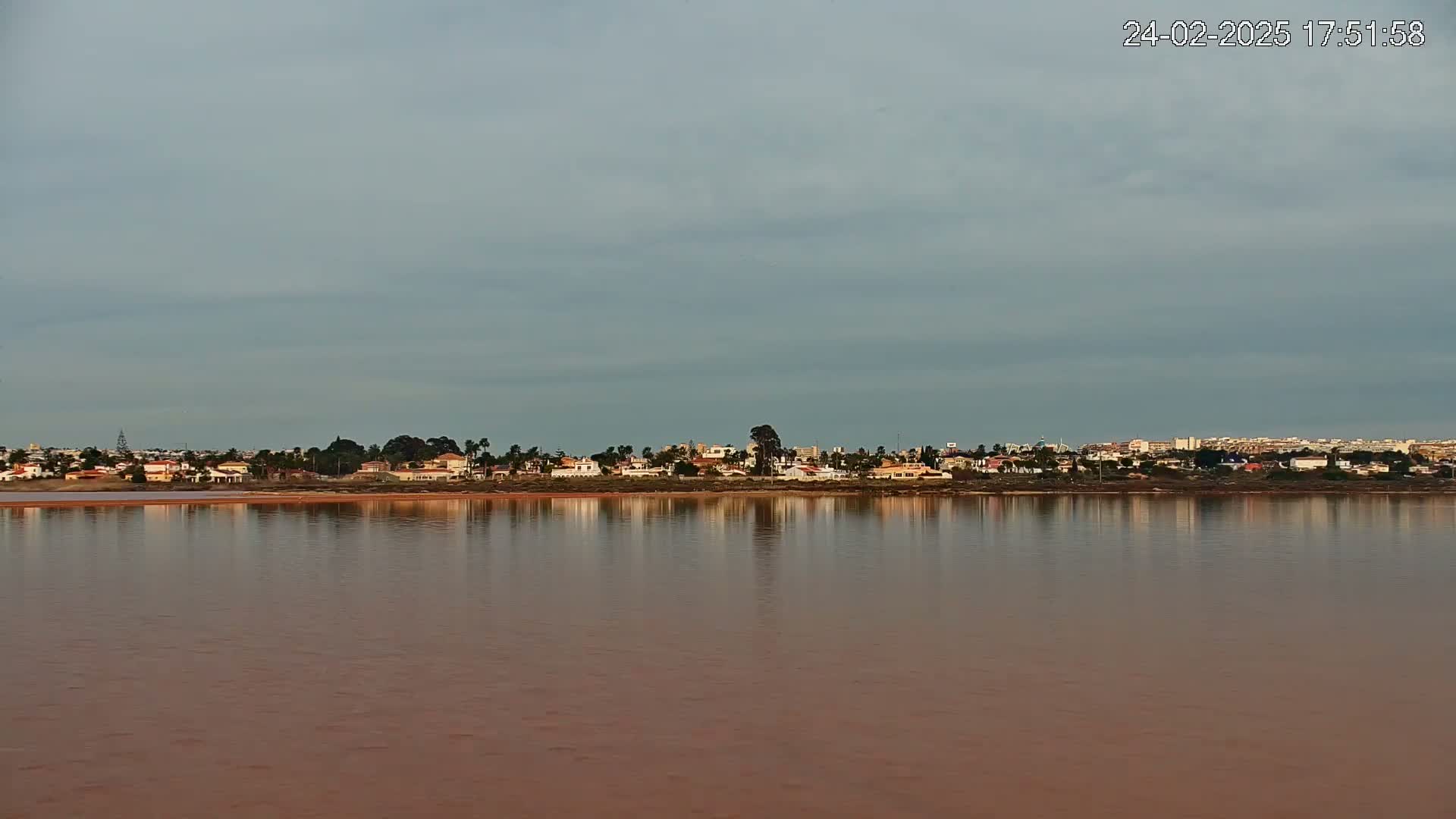 A calm body of reddish-brown water reflects a line of houses and trees under a cloudy sky.