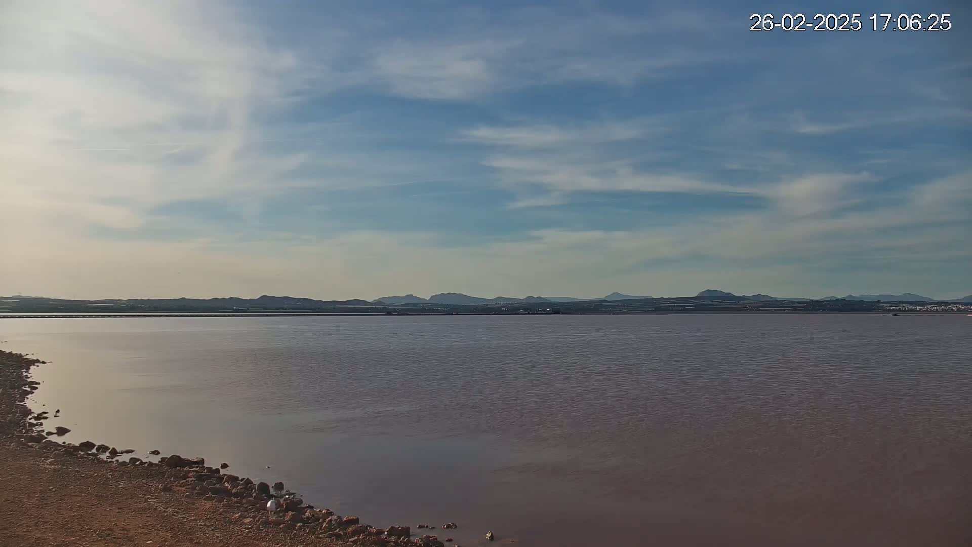 A calm body of pinkish-brown water stretches towards a distant mountain range under a partly cloudy sky.