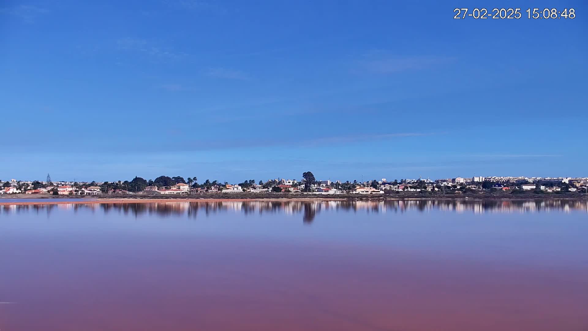A calm, pinkish lake reflects a clear blue sky and a distant town under sunny conditions.