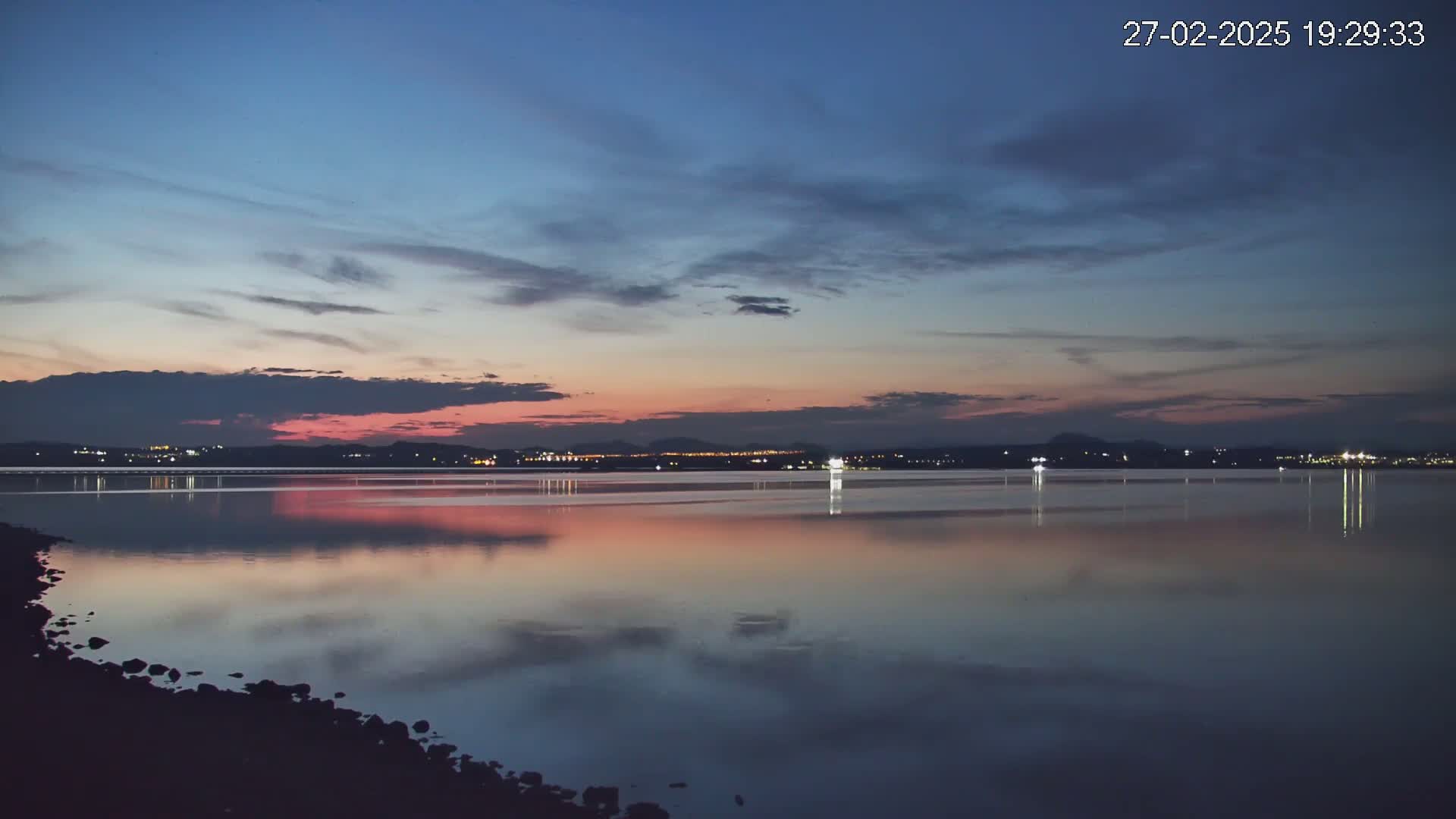 A calm body of water reflects a twilight sky with pink and purple hues over a distant city skyline.