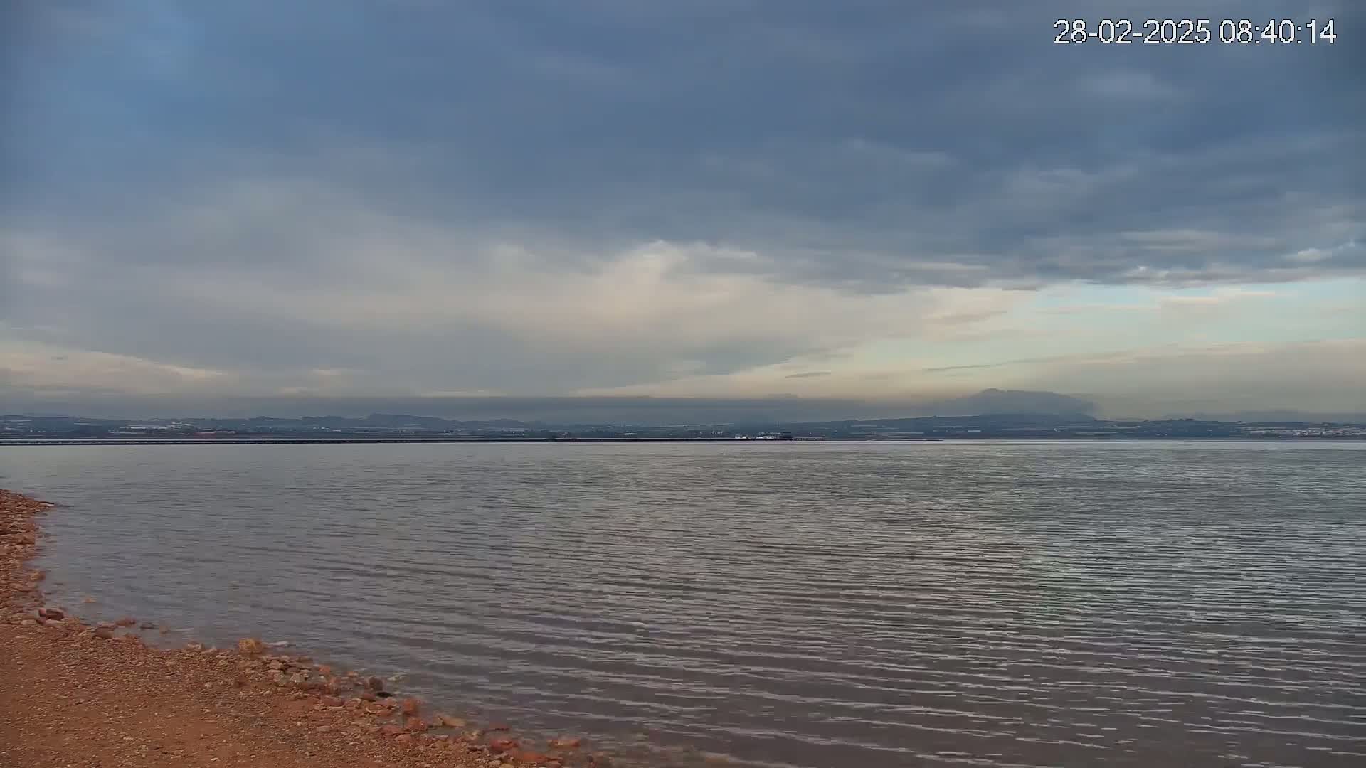 A calm body of water with a reddish-brown shoreline stretches towards a distant landmass under a mostly cloudy sky.
