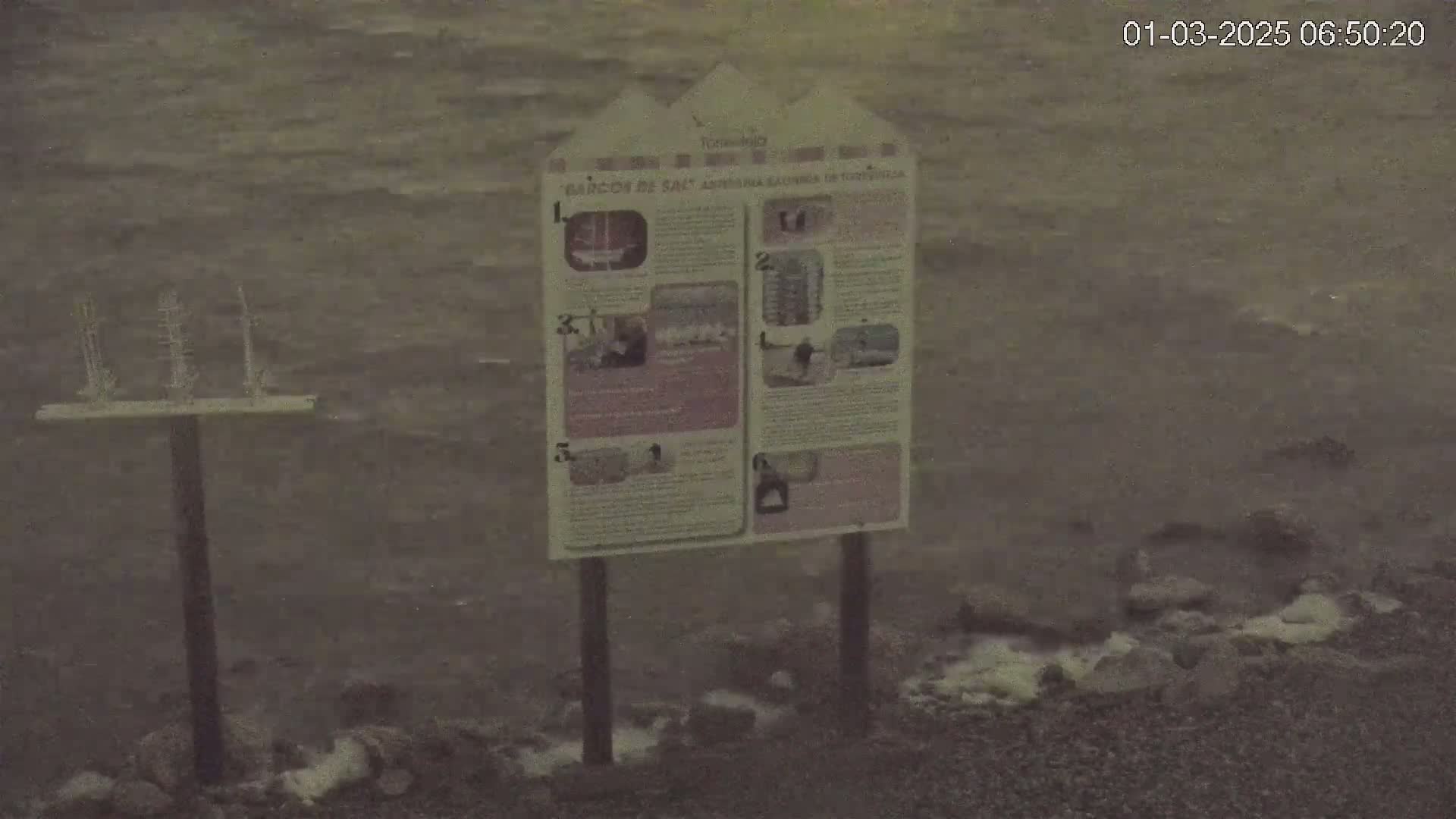 A model of a ship stands on a post near an informational sign by a choppy body of water.