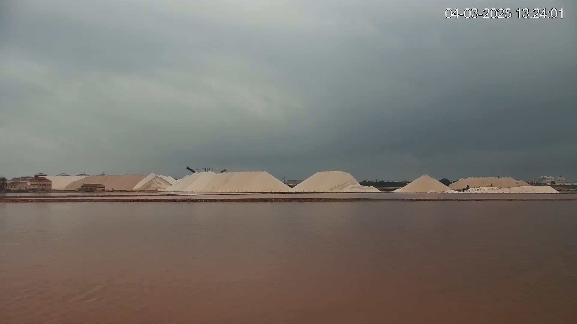 A body of reddish-brown water sits in front of several large piles of white salt under an overcast sky.
