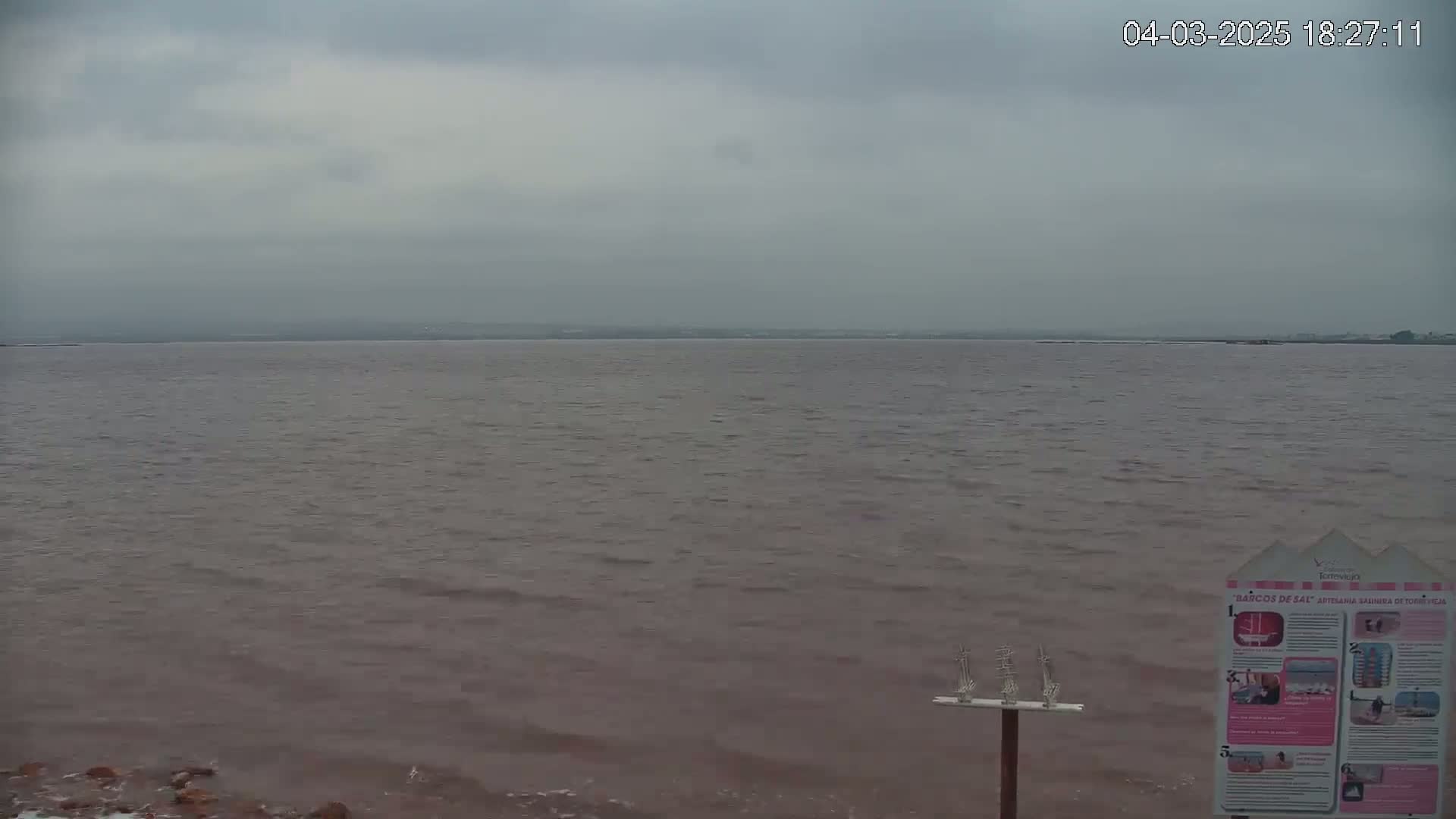 A calm, reddish-brown body of water stretches to a distant shoreline under an overcast sky.