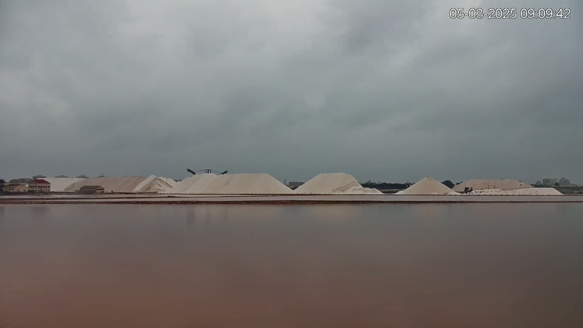 A body of still, reddish-brown water reflects a row of large, white salt piles under an overcast sky.
