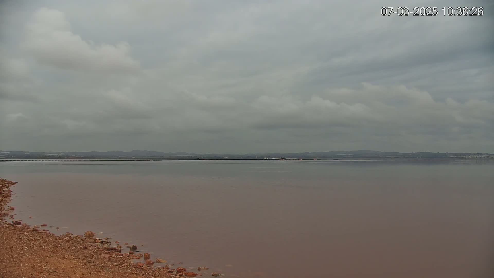 A calm, pinkish-brown body of water stretches to a distant shoreline under a cloudy sky.
