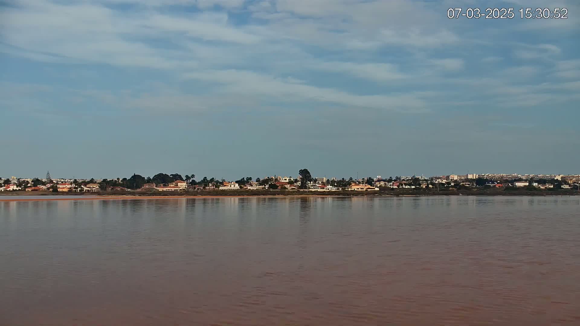 A calm, reddish-brown body of water reflects a shoreline of houses and vegetation under a mostly clear, lightly cloudy sky.
