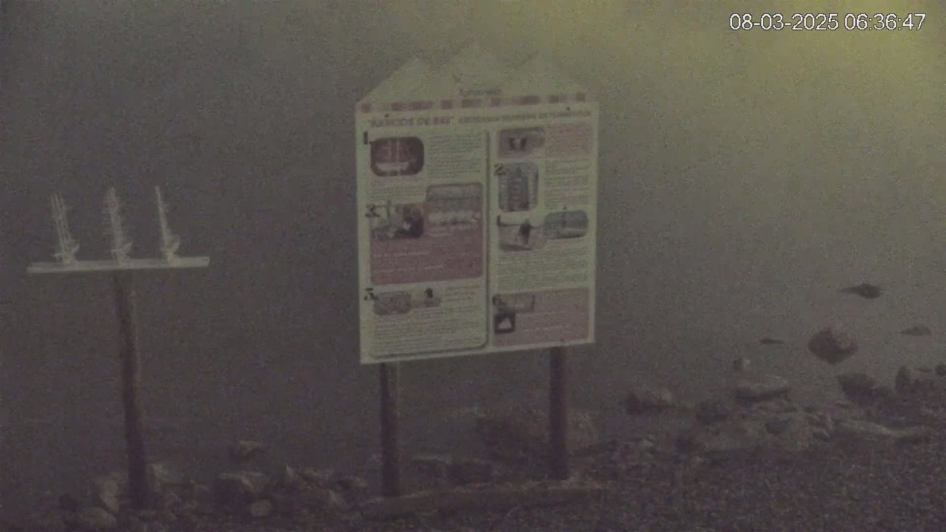 A sign stands near a post with three small model sailboats on top, all in a foggy or hazy outdoor environment.