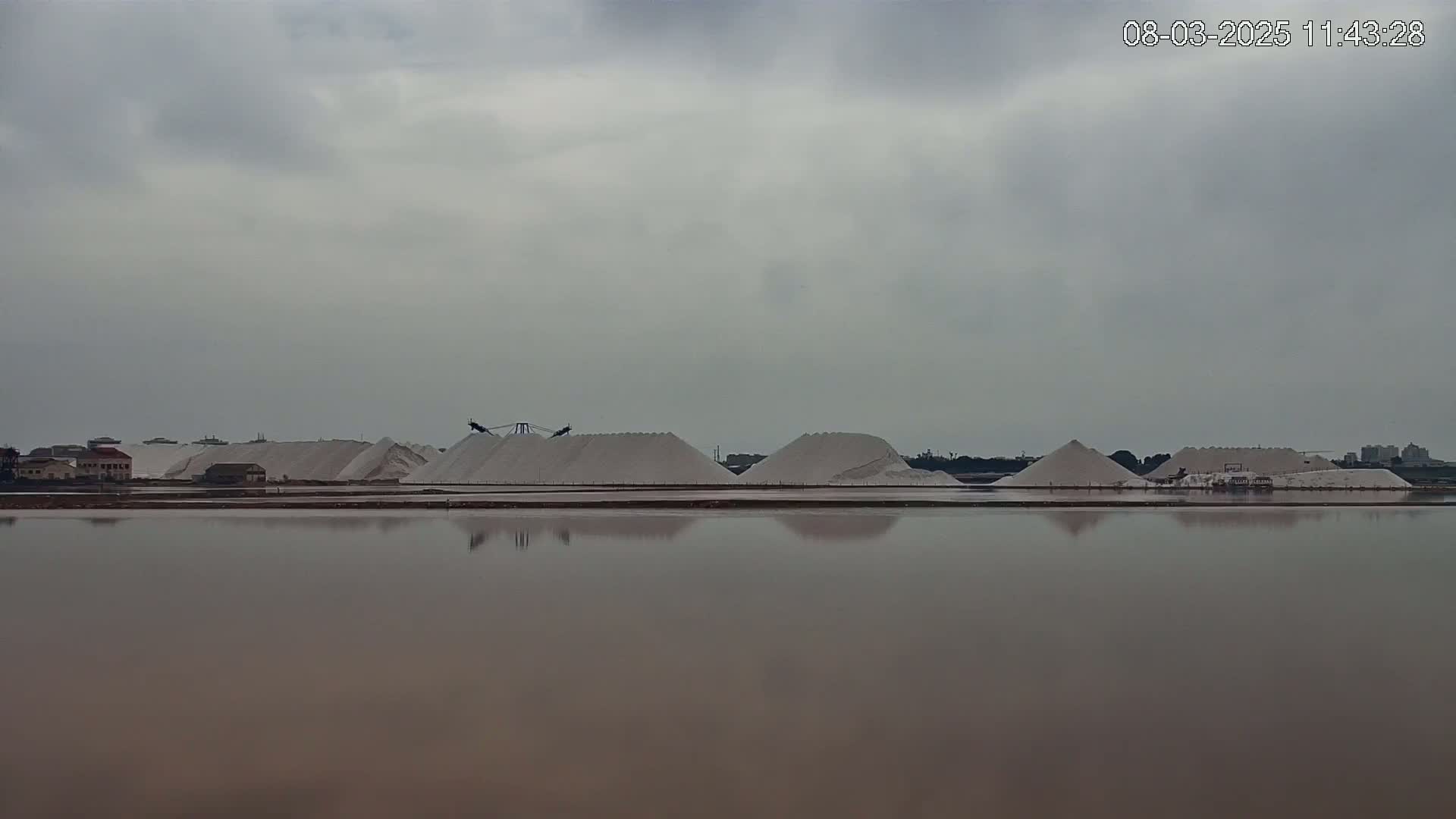 A large body of calm, brown water reflects a flat landscape of several large piles of white salt, industrial machinery, and a few buildings under an overcast sky.