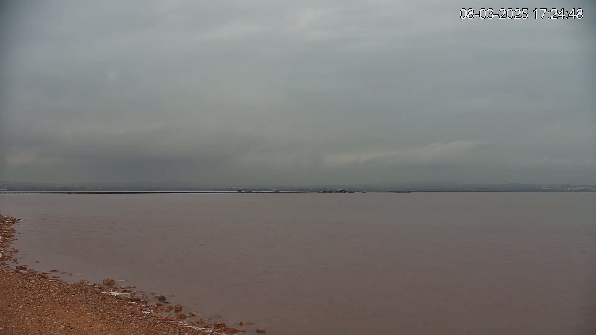 A pinkish-brown body of water stretches to a distant shoreline under an overcast sky.