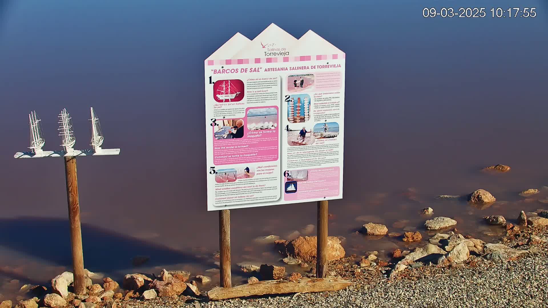 A pink-hued body of water, bordered by rocks and gravel, features a wooden post holding model sailboats and an informational sign.