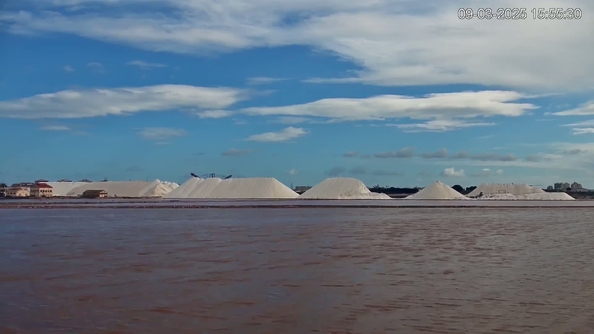 Several large piles of white salt are situated near a body of reddish-brown water under a partly cloudy blue sky.