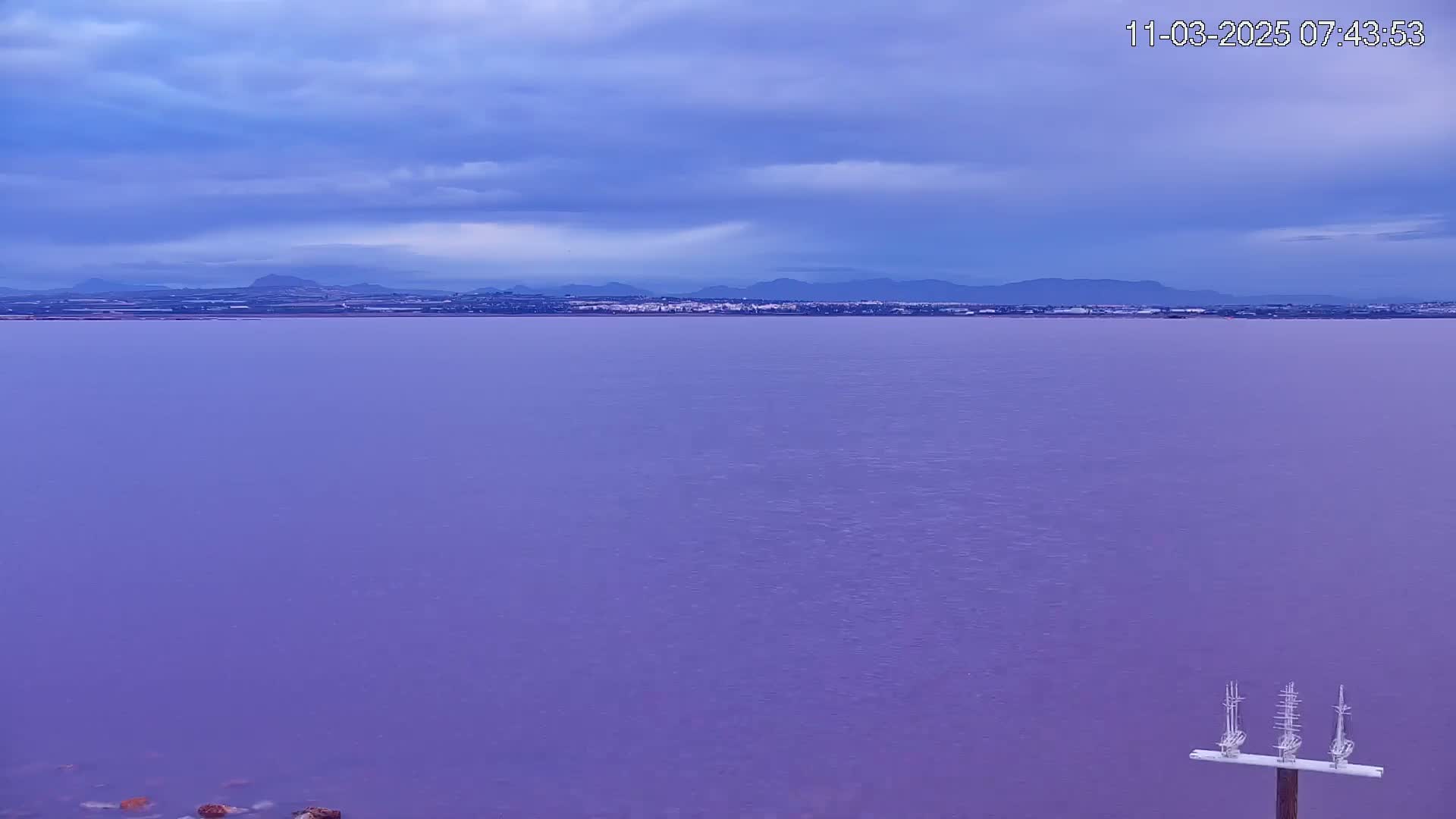 A calm, purplish body of water stretches to a distant shoreline with low mountains under a cloudy sky.