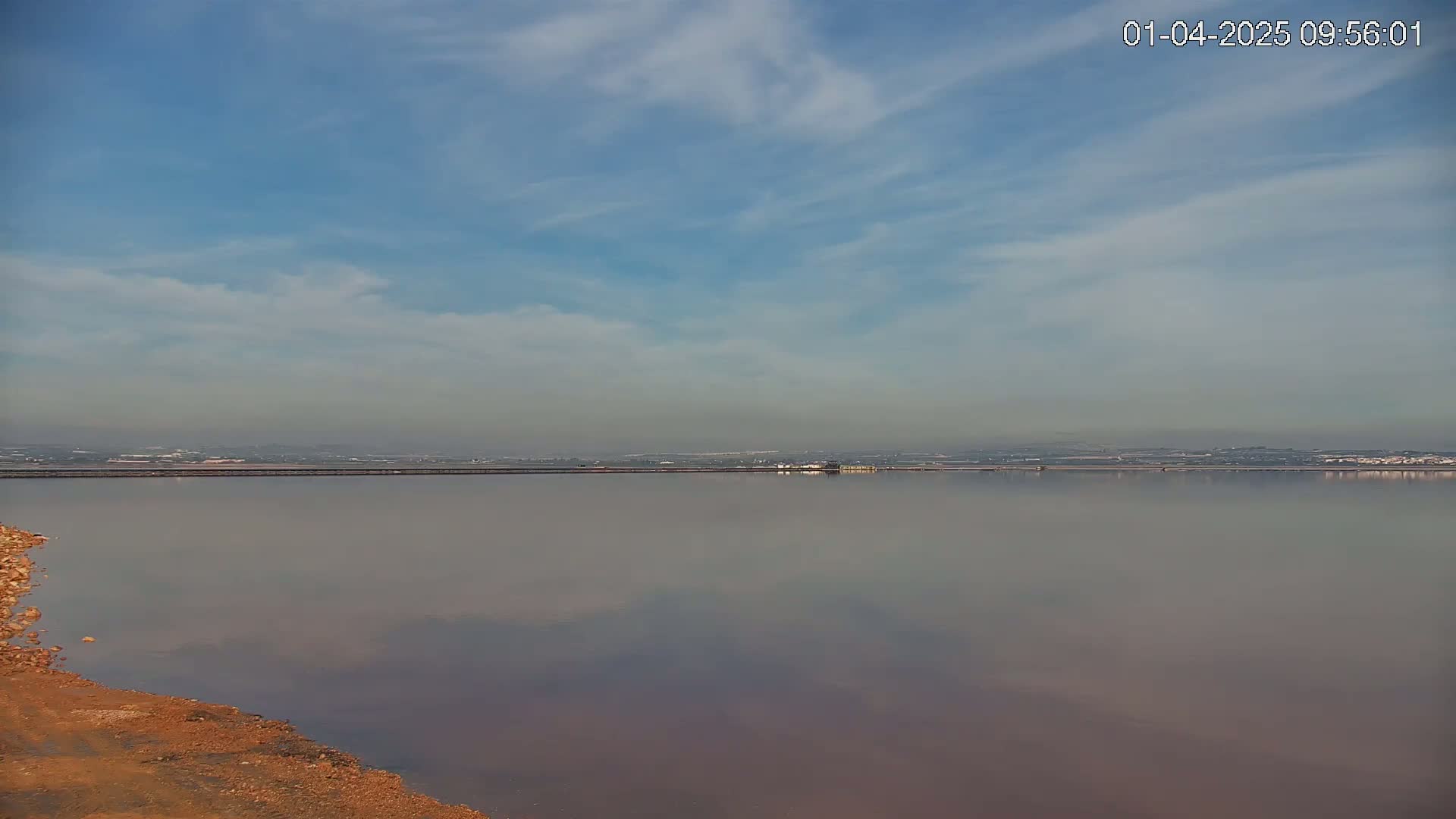 A calm, pink-hued salt flat reflects a hazy sky under a mostly clear, light-blue sky.