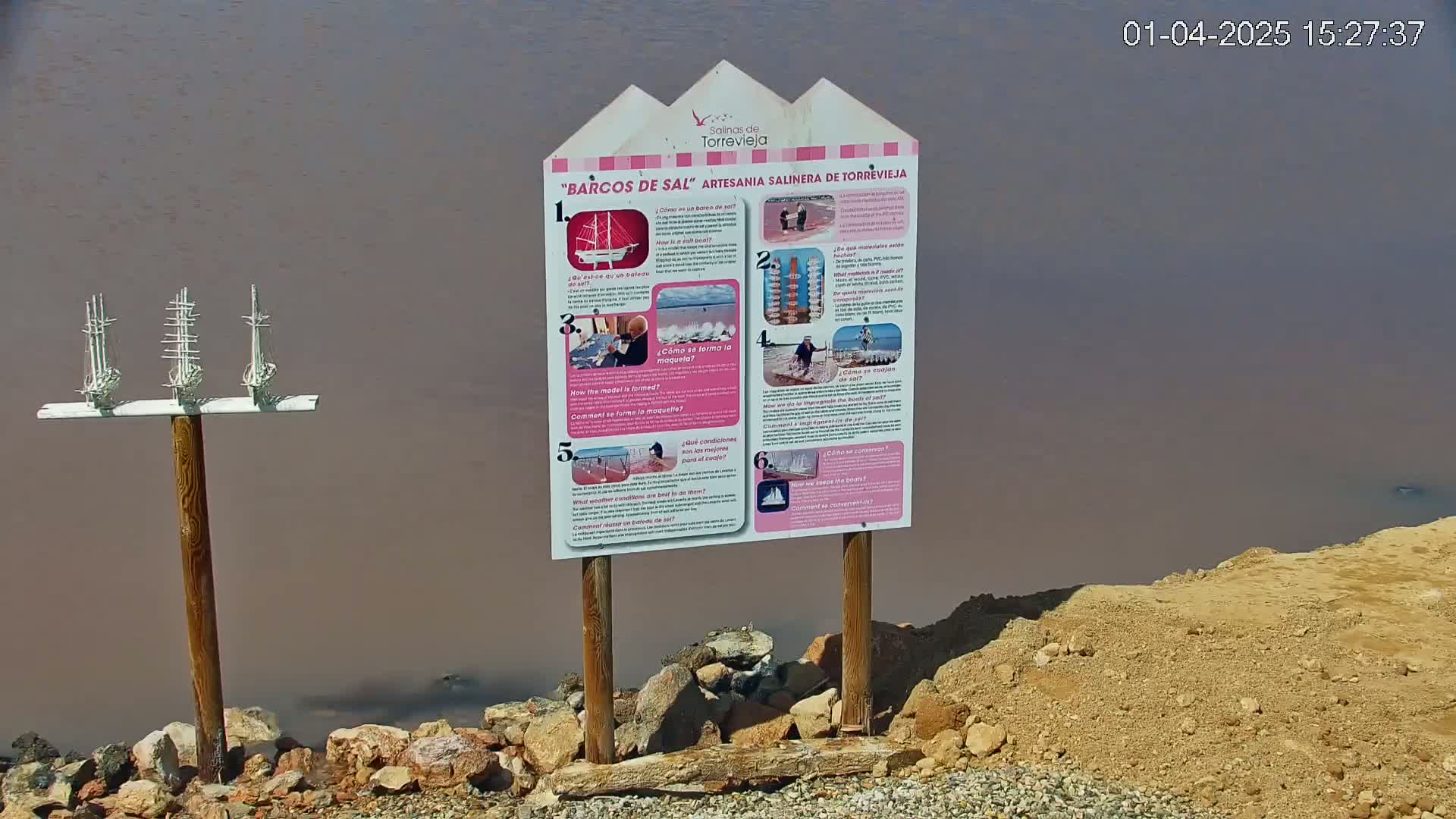 A wooden sign with informational panels about salt boats stands near a body of pinkish-brown water, next to a small wooden stand with three miniature salt boat sculptures.