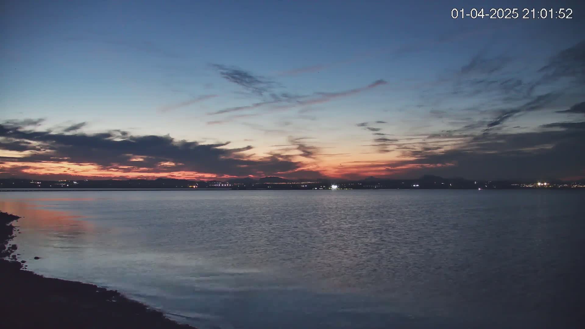 A calm body of water reflects a dusky sunset over a distant, city-lit shoreline under a partly cloudy sky.
