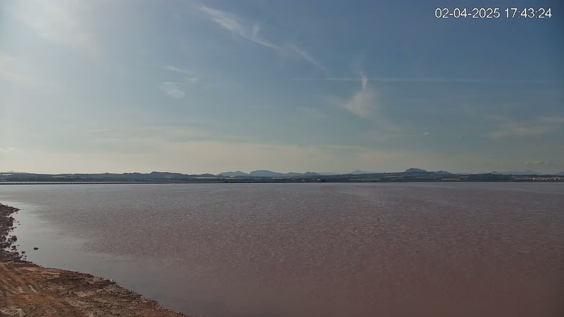 A pinkish-brown body of water stretches to a distant line of low hills under a mostly clear, sunny sky.