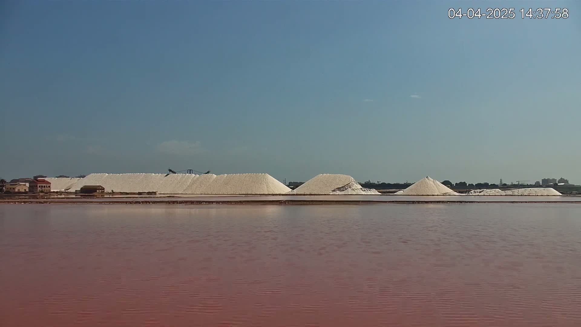Across a calm, pinkish-red body of water, several large piles of white salt sit on the shore under a clear blue sky.