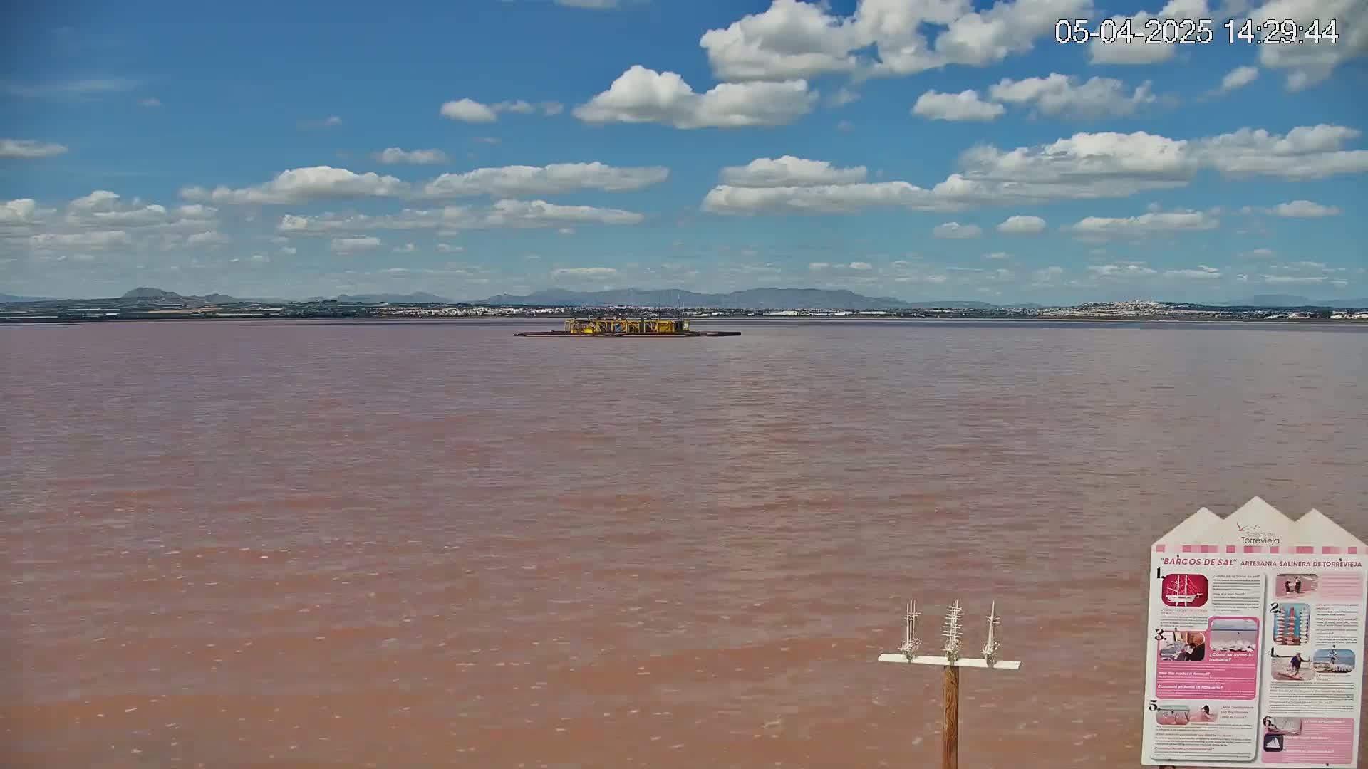 A pinkish-red body of water stretches to a distant shoreline under a partly cloudy blue sky, with a small structure visible in the middle distance.