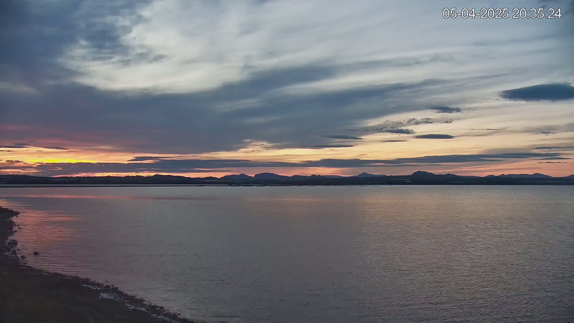 A calm body of water reflects a sunset with pastel pink and orange hues under a mostly cloudy sky,  with a distant mountain range silhouetted against the horizon.
