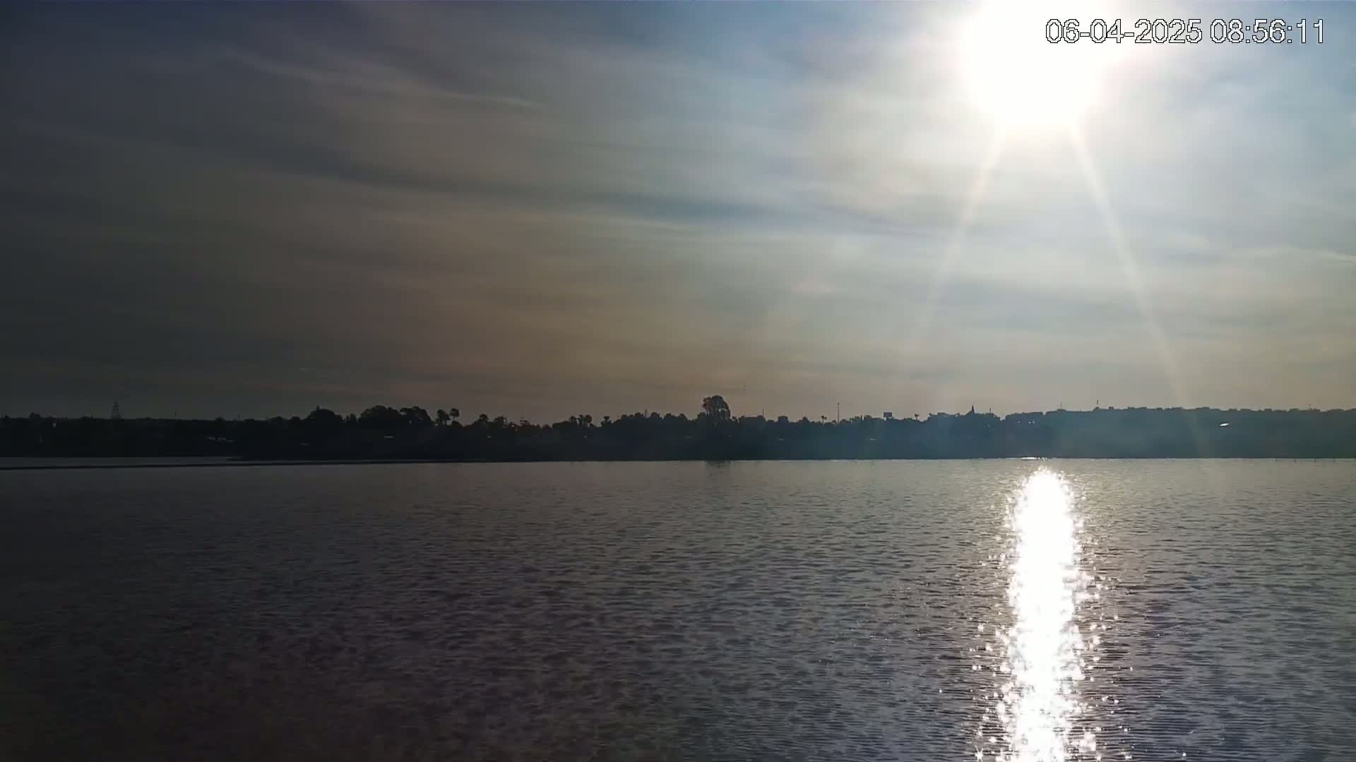 The sun shines brightly over a calm body of water, with a distant shoreline of trees and buildings silhouetted against the sky.