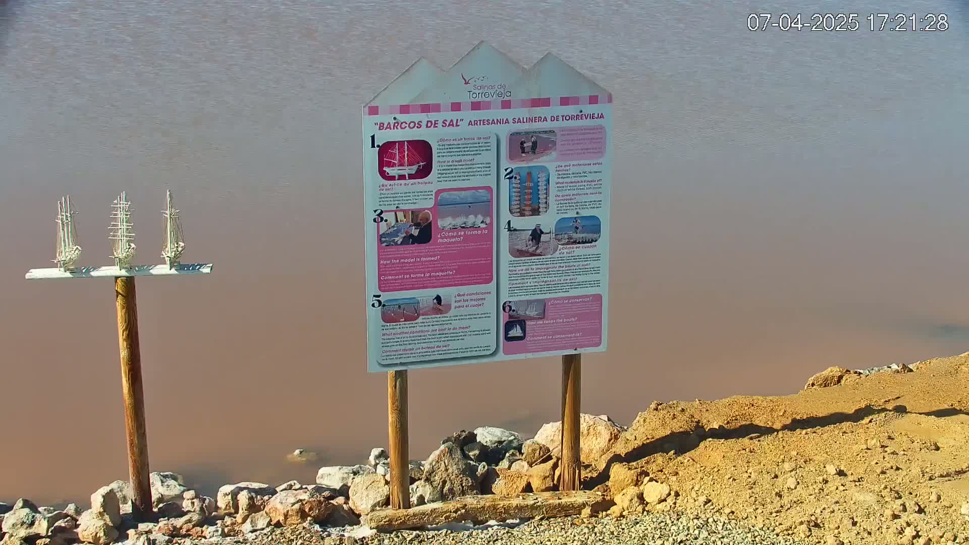 A wooden post holding three small model sailboats stands near an informational sign by a body of pinkish-brown water.