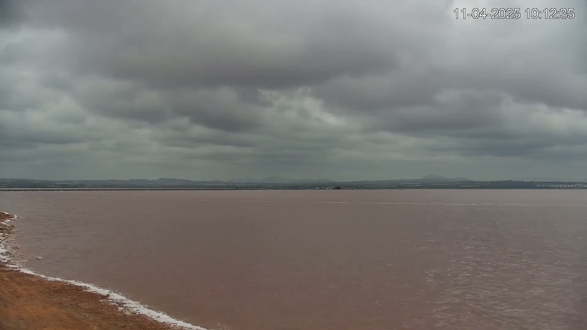 A pinkish-brown body of water stretches to a distant, low-lying shoreline under a cloudy, overcast sky.