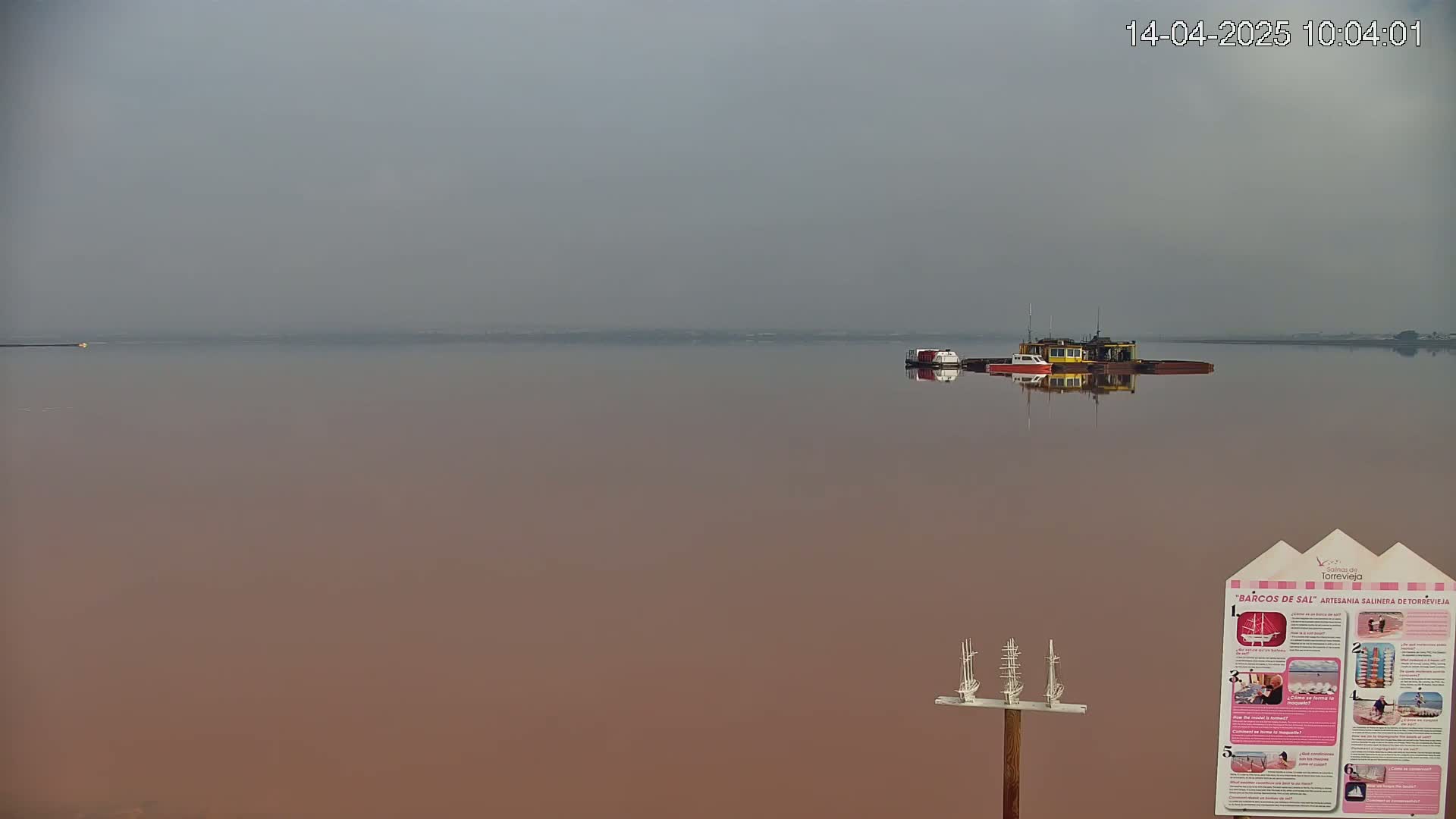 A calm, hazy body of pinkish water contains several small boats, one of which is noticeably larger than the others.