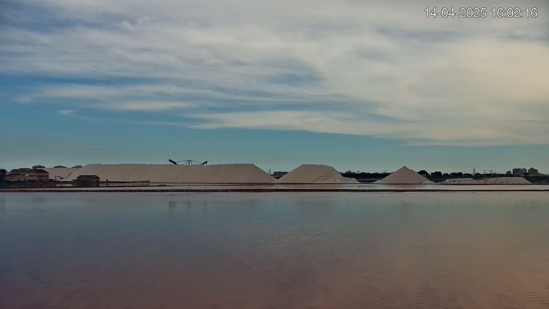 Large piles of white salt sit next to a calm body of water under a partly cloudy sky.