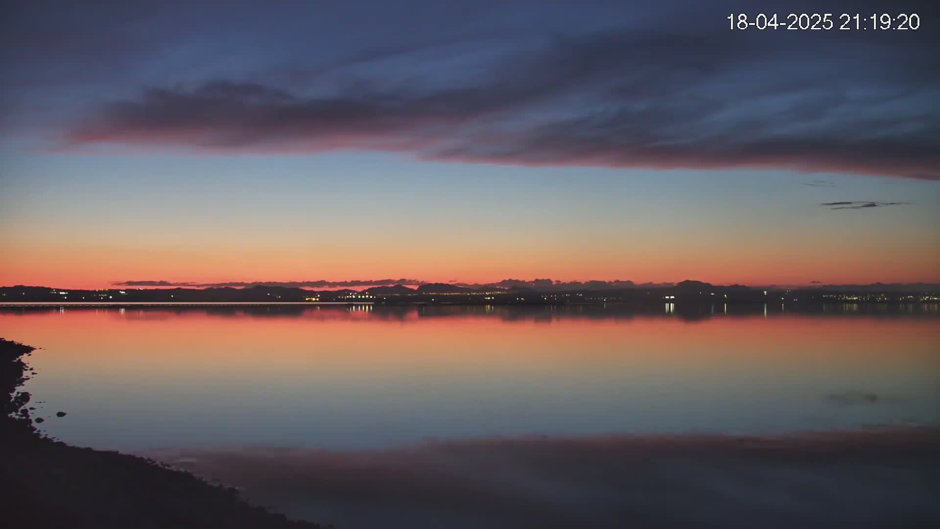 A calm body of water reflects a pink and orange sunset under a mostly cloudy sky.
