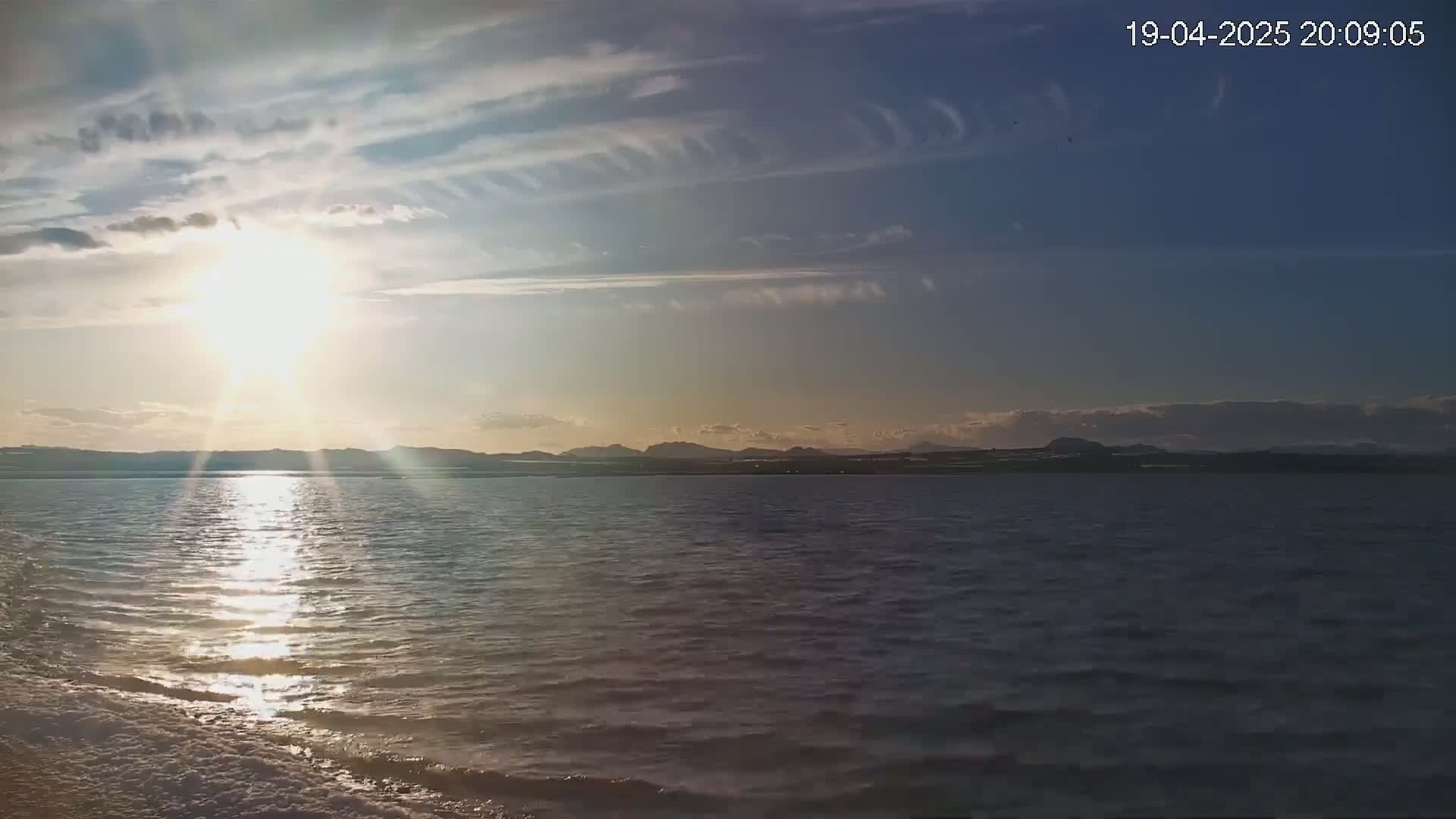A bright sun shines over calm water with a distant mountain range visible under a partly cloudy sky.