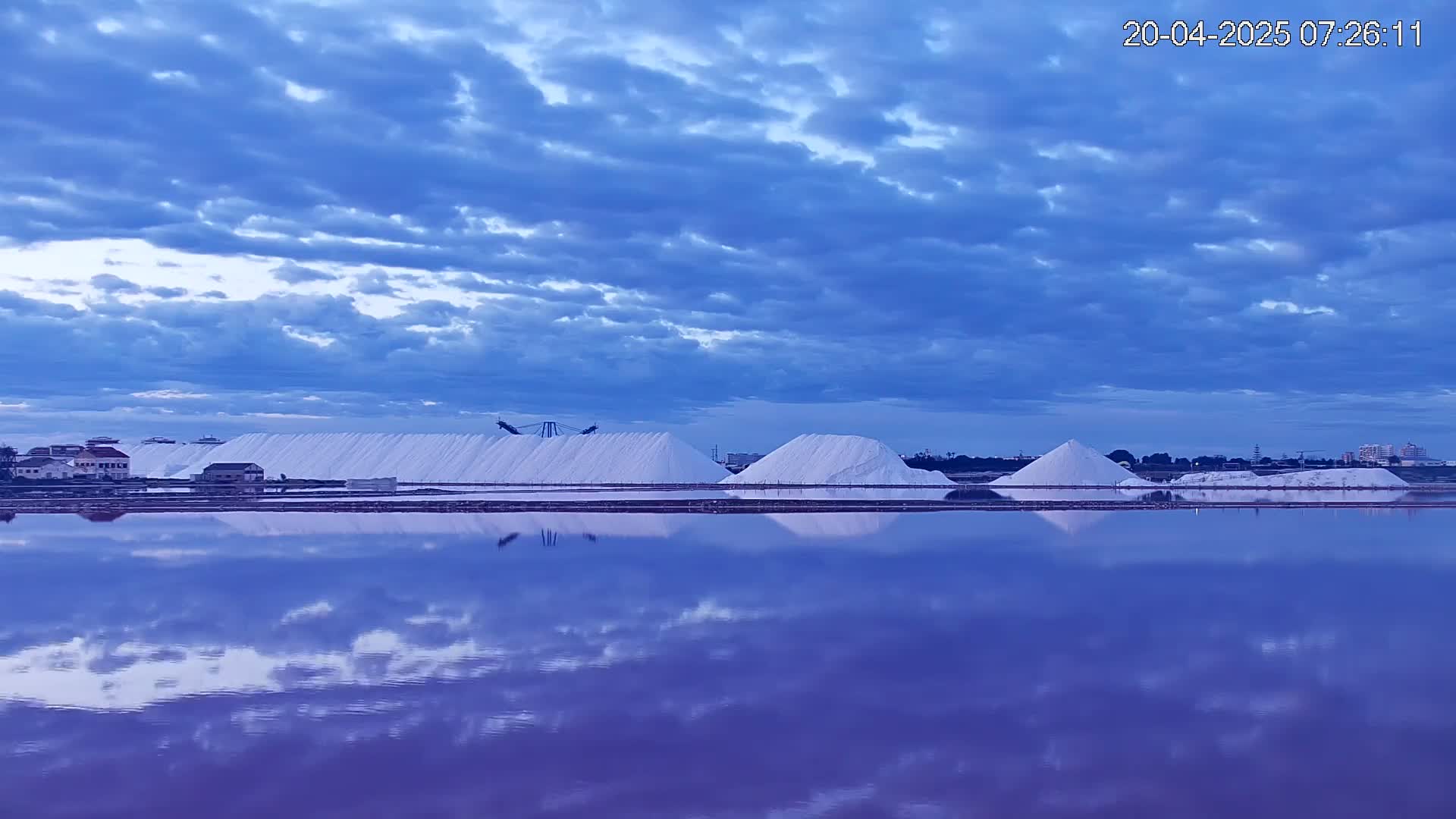 Large piles of white salt are reflected in a calm, purplish-blue body of water under a partly cloudy blue sky.