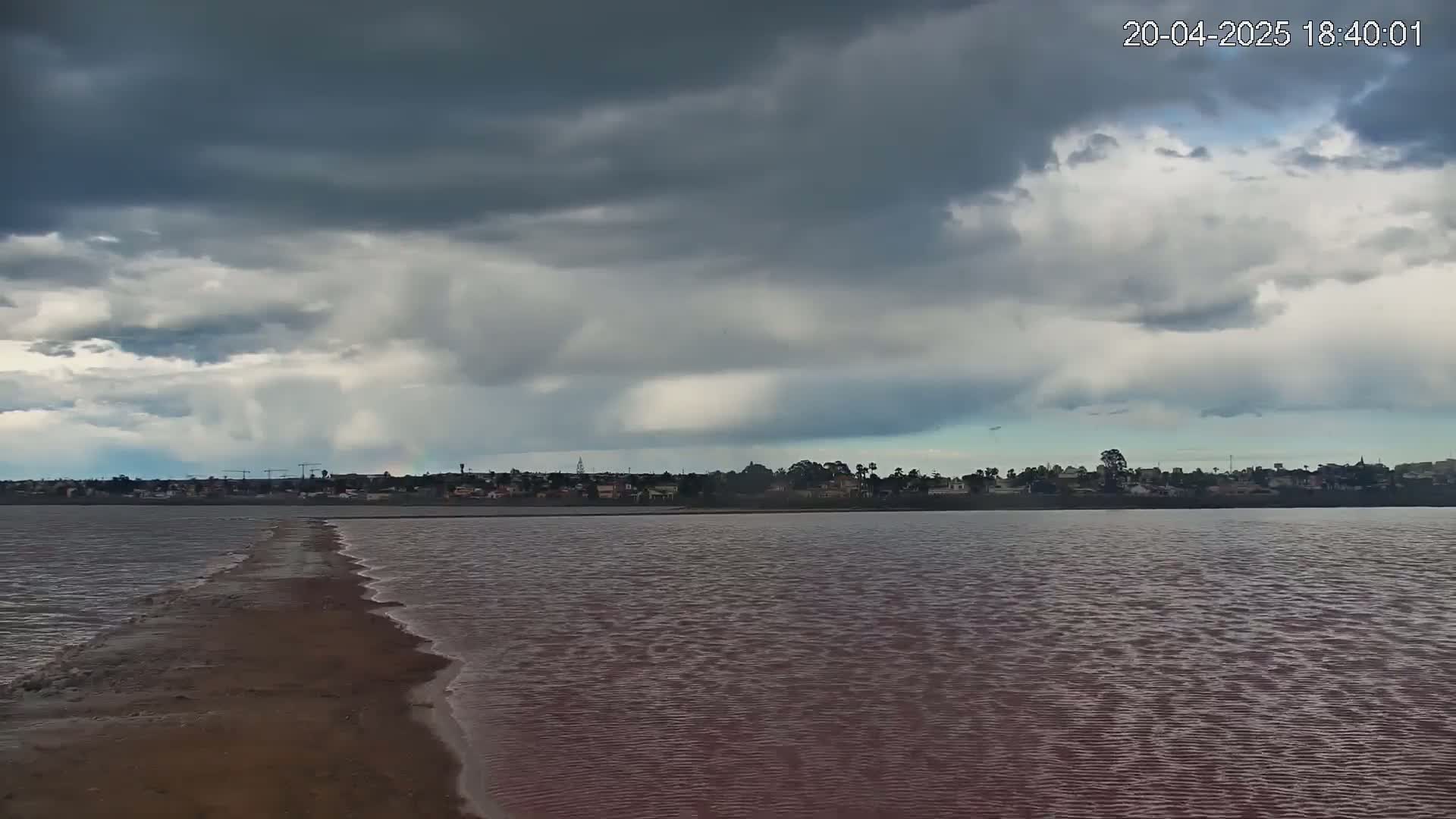 A narrow, land bridge extends across a pinkish-red body of water under a mostly cloudy, overcast sky.