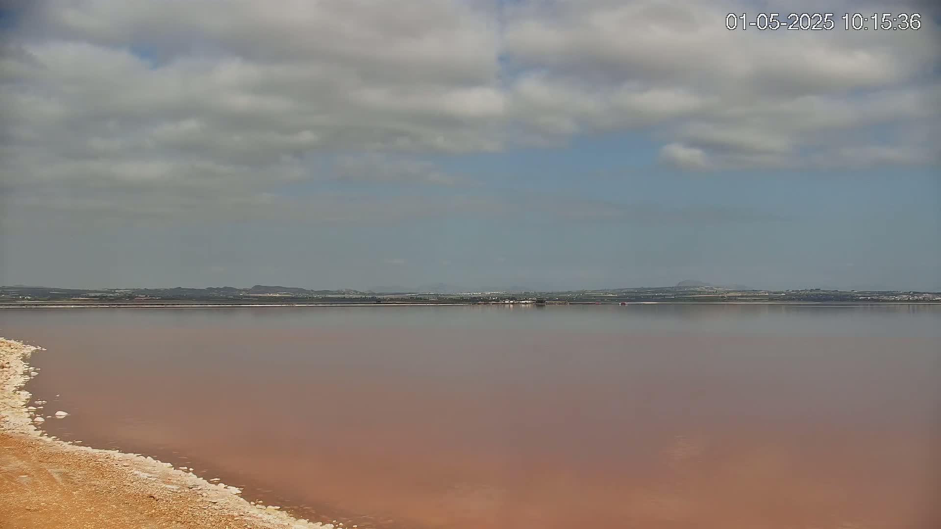 A pinkish-red lake stretches to a distant shoreline under a partly cloudy sky.