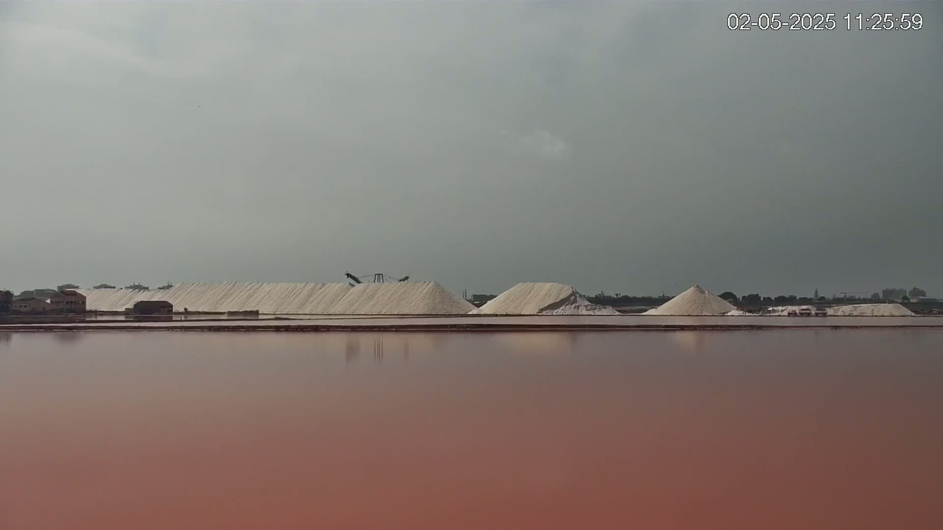 Several large piles of white salt are situated on the far shore of a pinkish-red body of water under a cloudy sky.