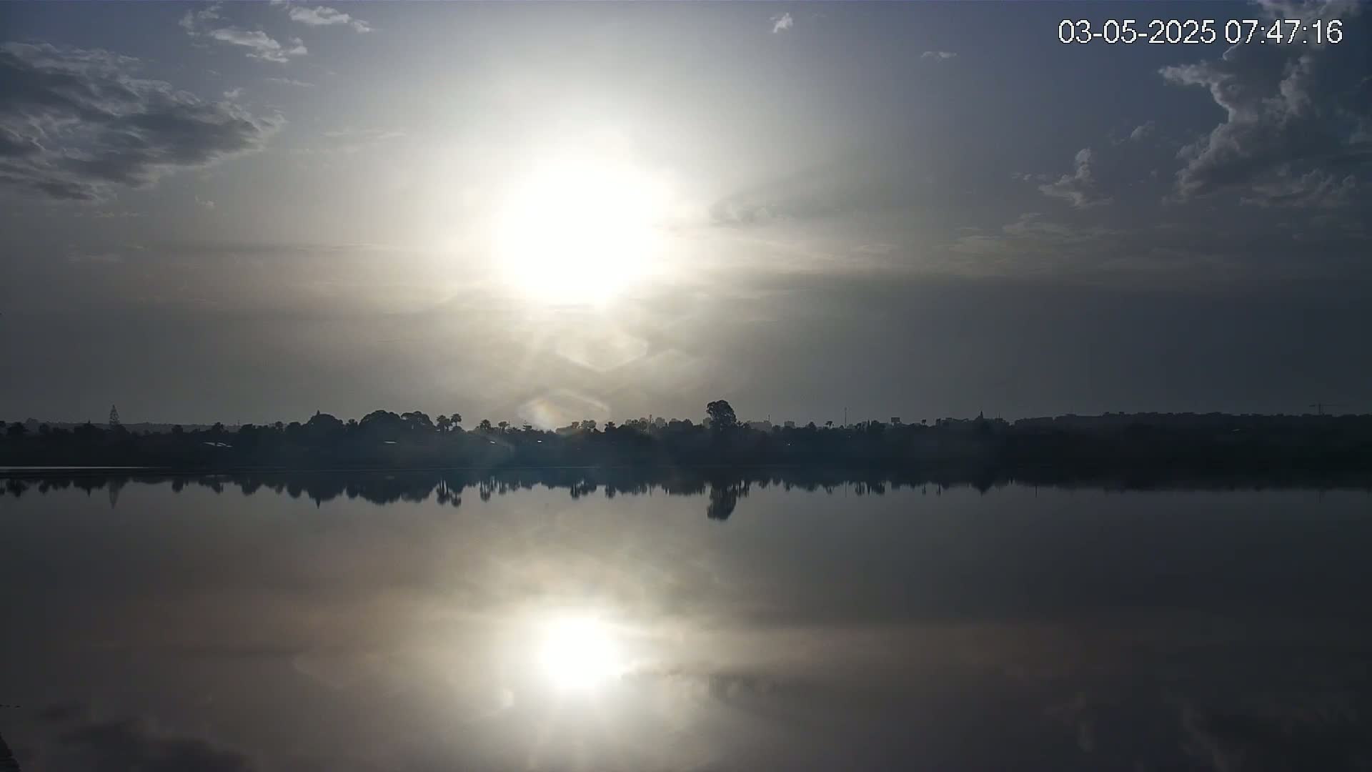 A calm lake reflects a bright sunrise under a partly cloudy sky.