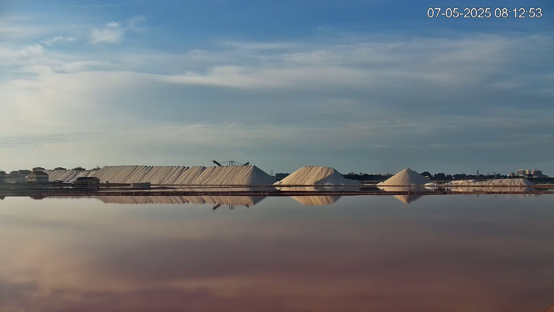 Several large piles of white salt are reflected in a calm, pinkish-red body of water under a mostly clear sky.