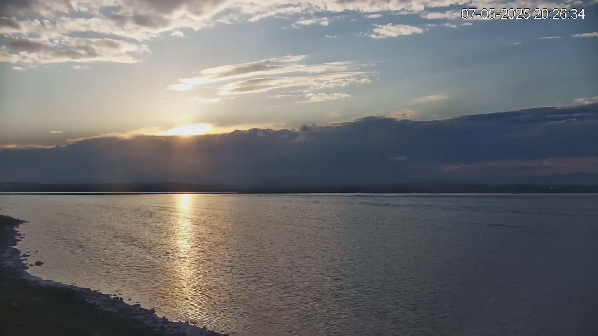 A calm body of water reflects the setting sun under a partly cloudy sky.