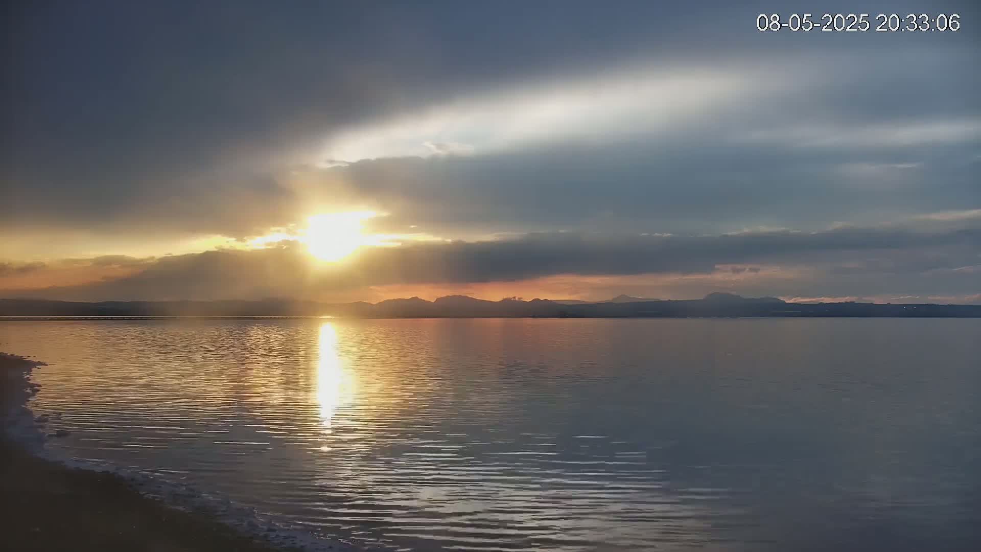 A calm body of water reflects the setting sun's rays under a partly cloudy sky, with distant mountains visible on the horizon.