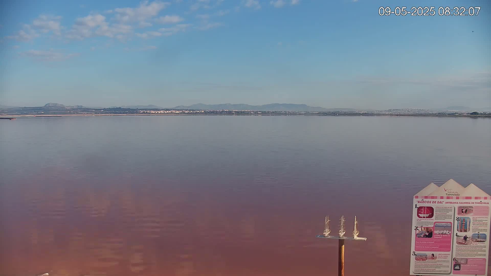 A calm, pinkish lake stretches to a distant shoreline under a mostly clear, light-blue sky.