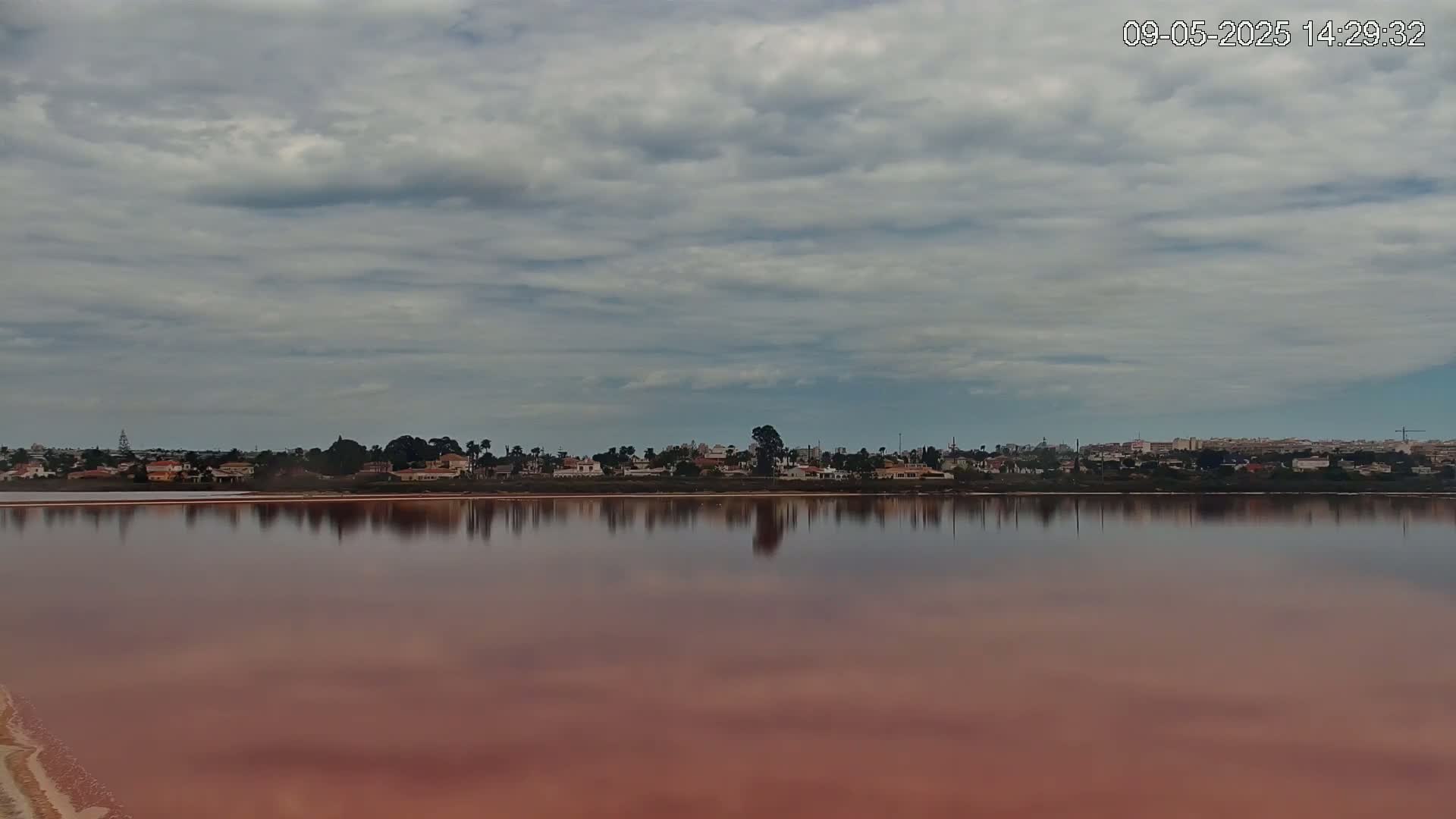 A calm, pinkish-red lake reflects a line of houses and trees under a cloudy sky.