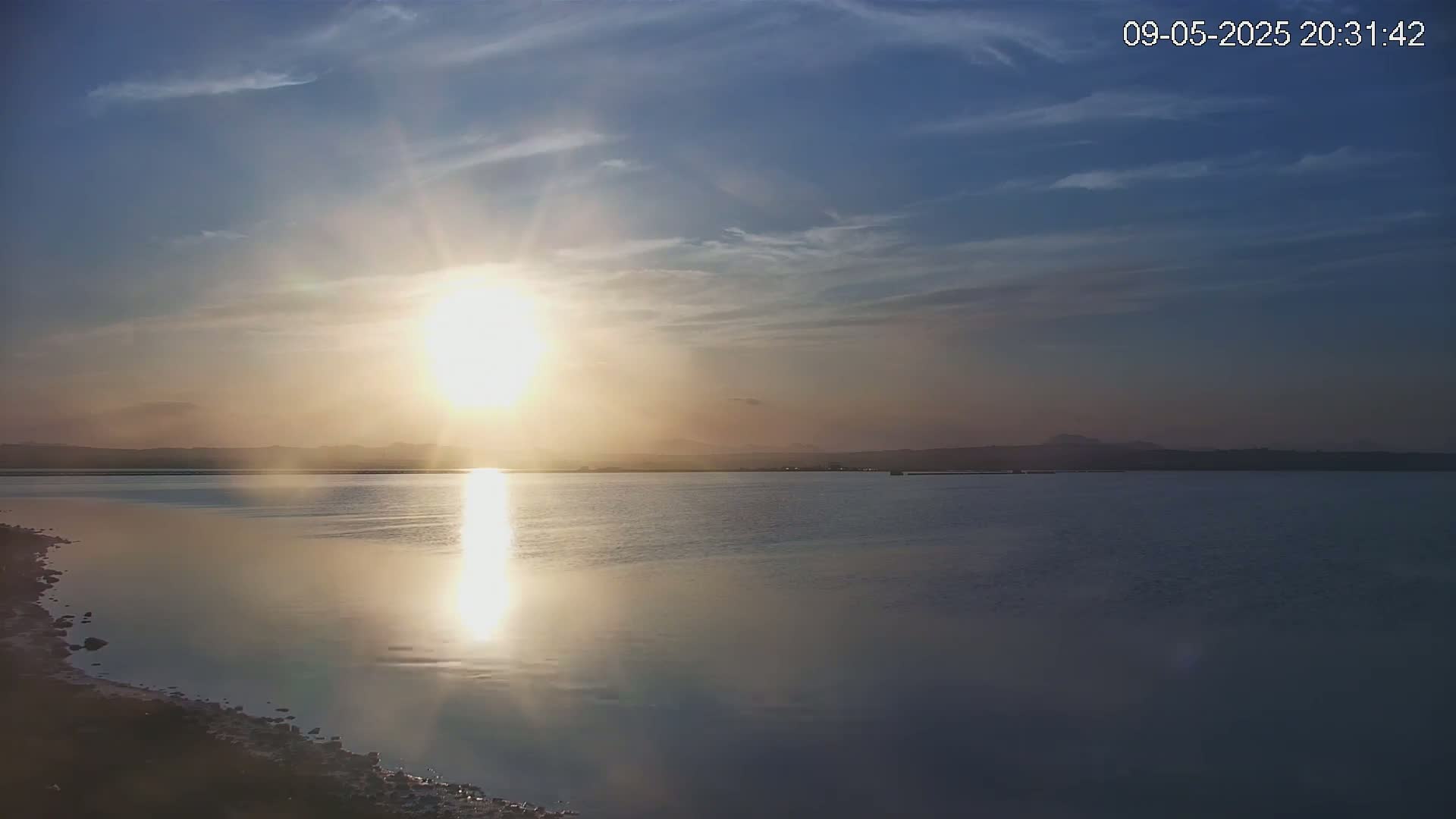A bright sun sets over a calm body of water, with a distant shoreline visible under a partly cloudy sky.