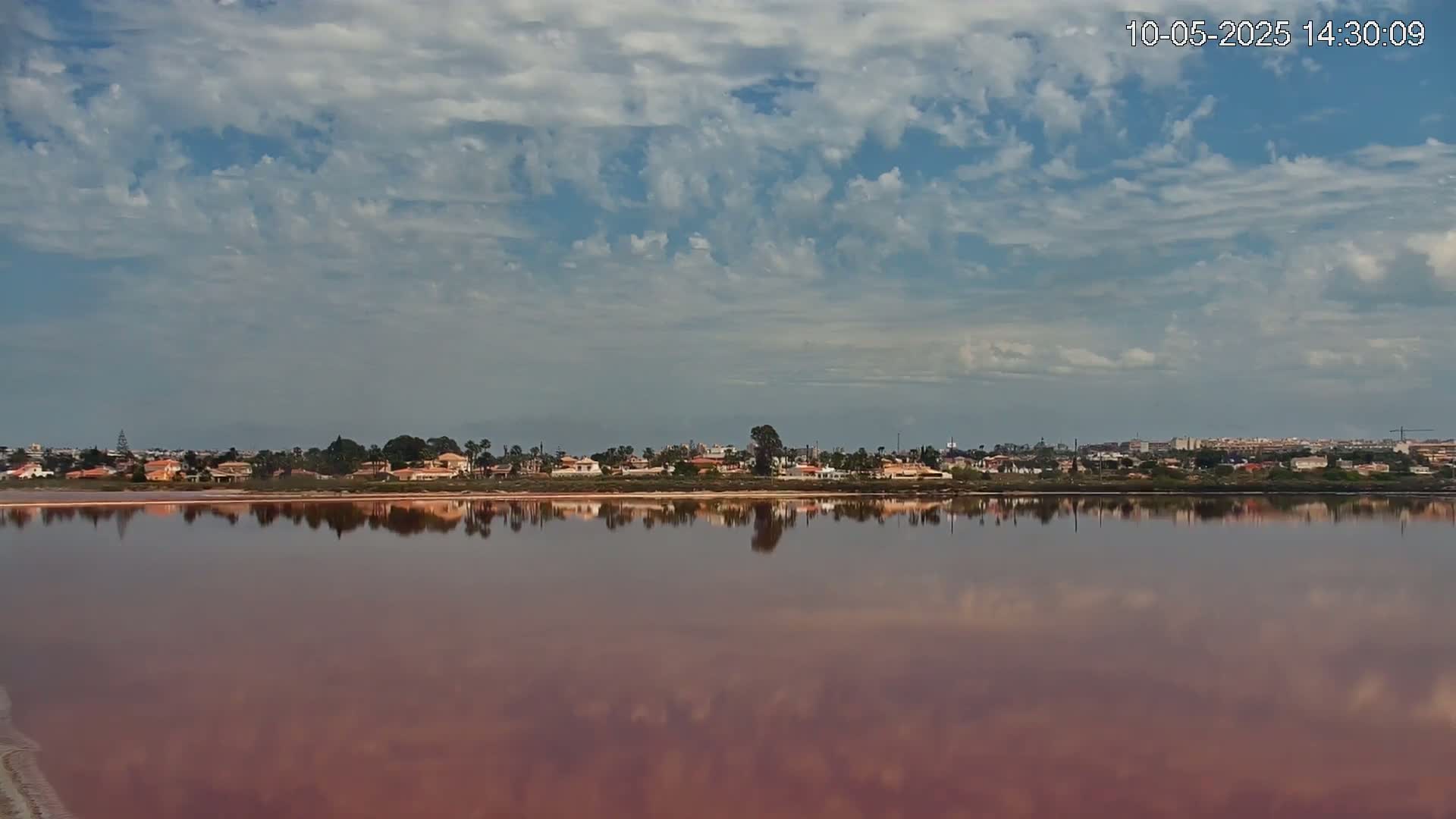 A pink lake reflects a residential area under a partly cloudy sky.