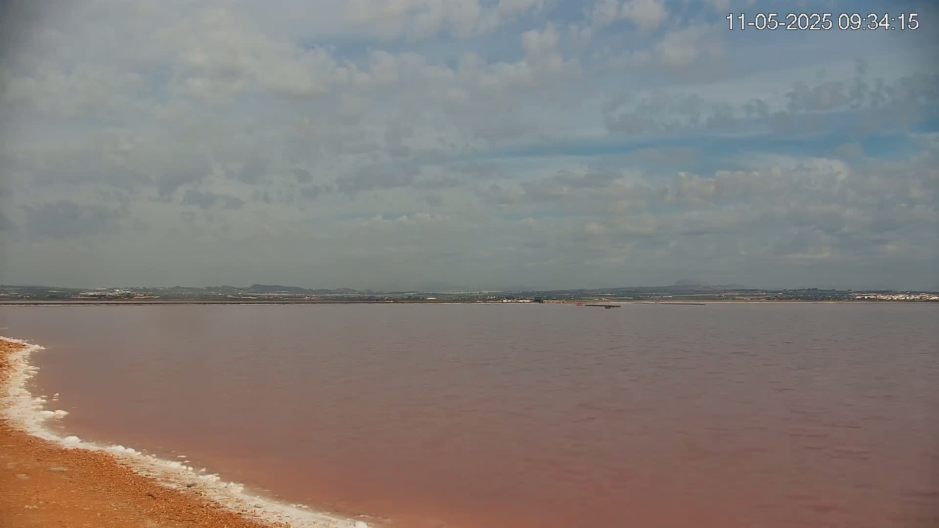 A pinkish-red body of water stretches to a distant shoreline under a mostly cloudy sky.